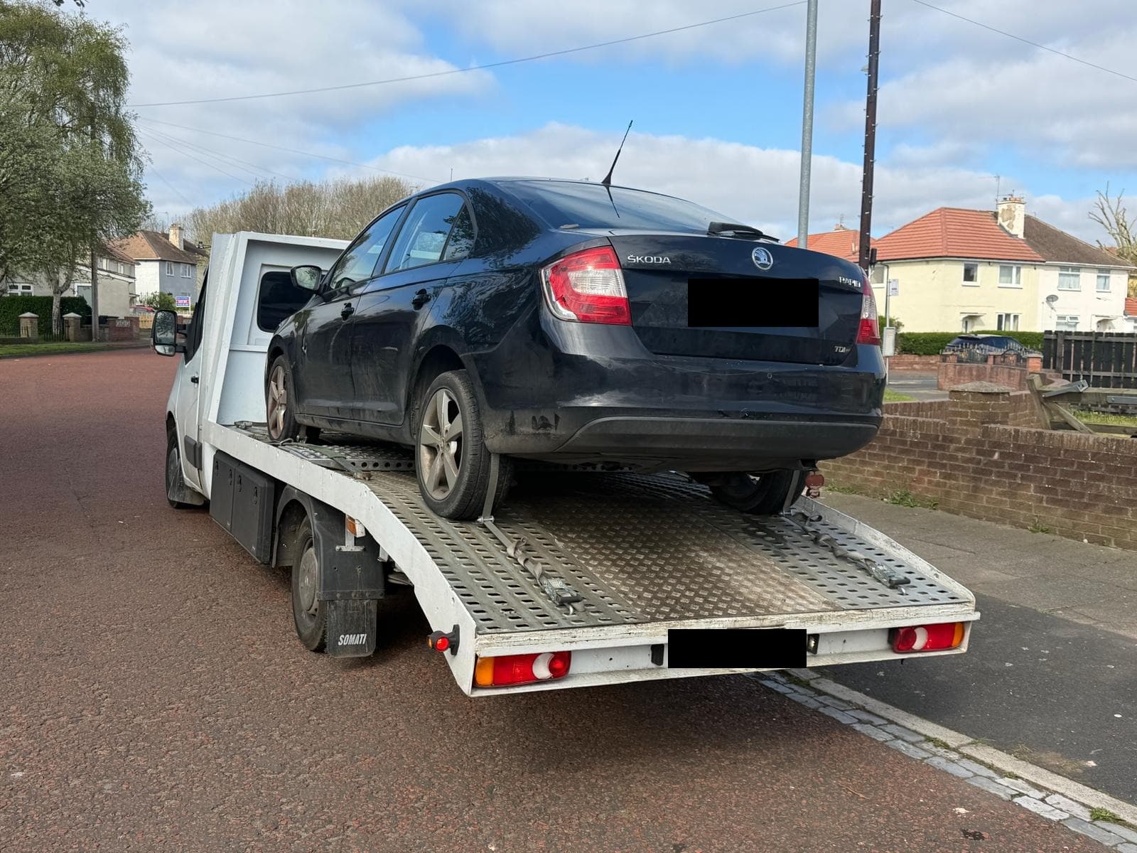 A dark blue Skoda Rapid on the back of a flatbed recovery truck on a residential street of semi-detached houses