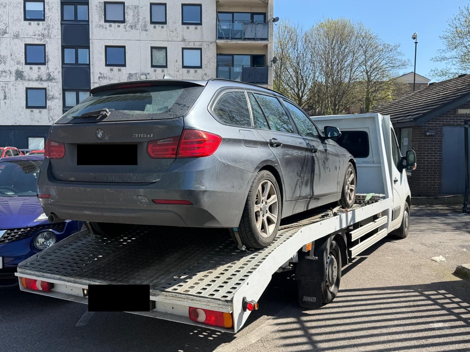 A grey BMW 3 Series estate loaded on a flatbed recovery truck in a car park, with a block of flats behind